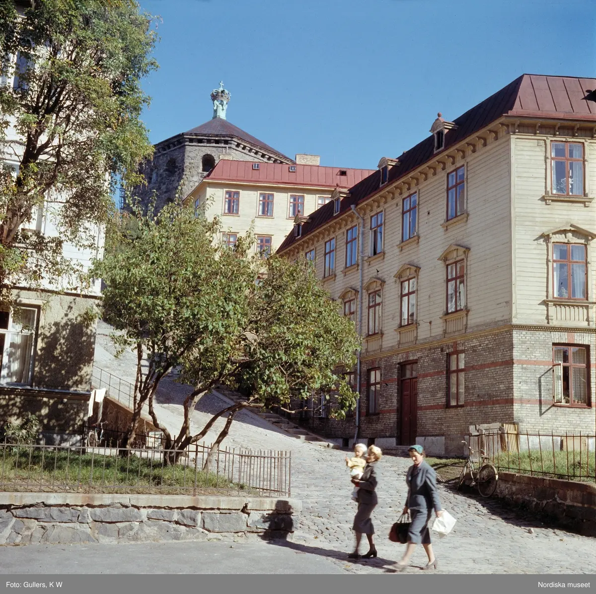 Risåsgatan 15-17 i Göteborg. Trähus, kullerstensgata. Kvinnor och ett barn i förgrunden. Skansen Kronan.