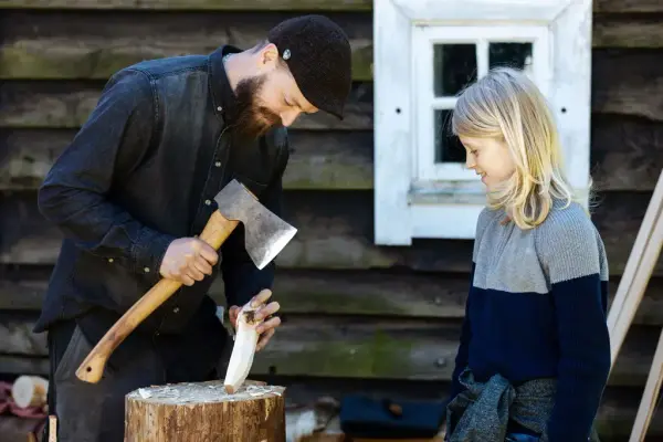 Mann med øks i handa som arbeider med tre, eit barn som ser på