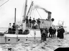 Ångfartyget Södra Wettern ligger vid kaj, i Gränna hamn? Män och kvinnor på kaj och ombord. En ung man keps sitter på en stol vänd mot fotografen och spelar dragspel.