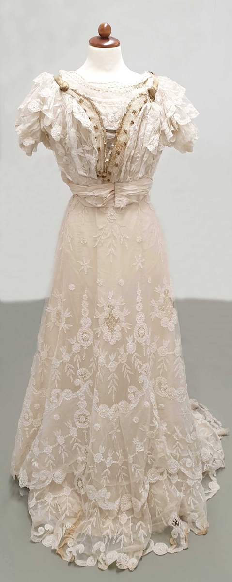 Kjolen av to deler - skjørt med påkneppet bluse.
Blusen er i organza og tyll med formede rosetter av blonder i metervis, påsydd en og en. Et bredt gullbånd med knute i enden rundt halsutringing. Blusen har korte puffermer, med blonder. Skjørtet har slep og består av tyll med broderte rosetter.