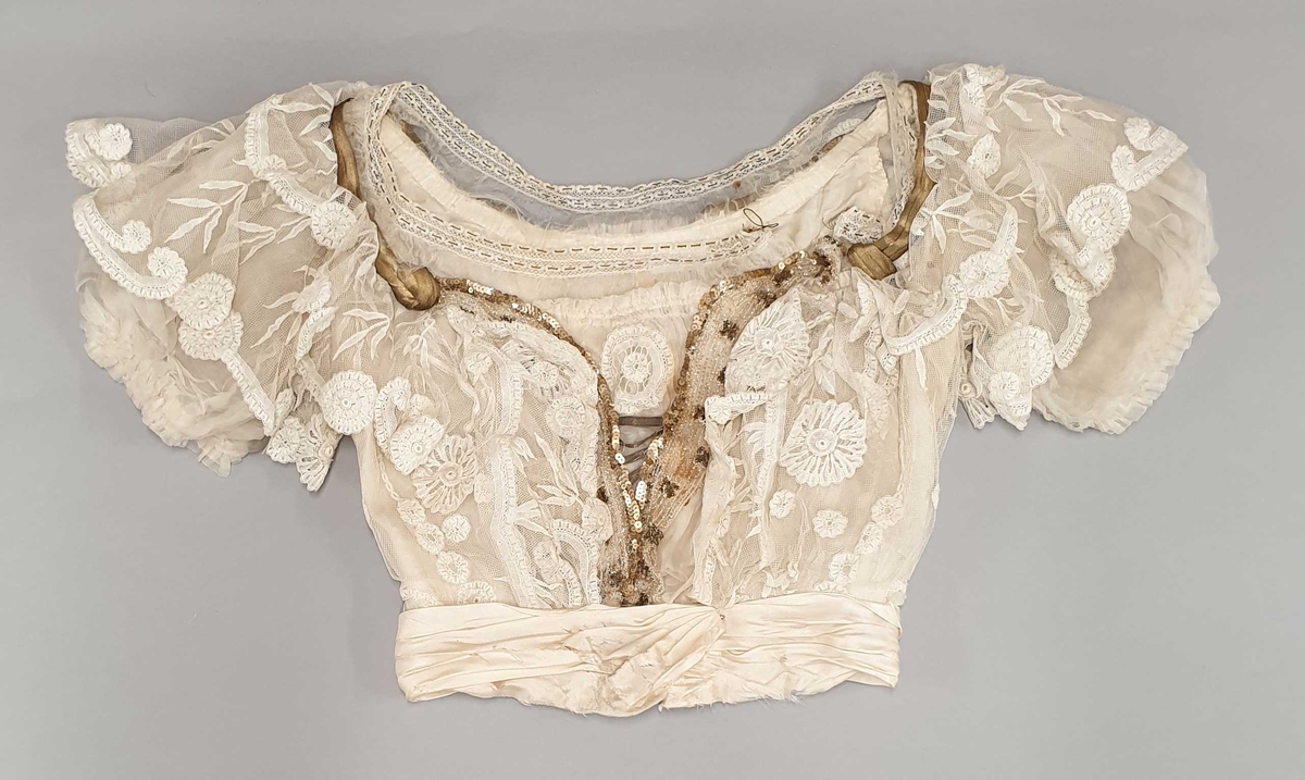 Kjolen av to deler - skjørt med påkneppet bluse.
Blusen er i organza og tyll med formede rosetter av blonder i metervis, påsydd en og en. Et bredt gullbånd med knute i enden rundt halsutringing. Blusen har korte puffermer, med blonder. Skjørtet har slep og består av tyll med broderte rosetter.