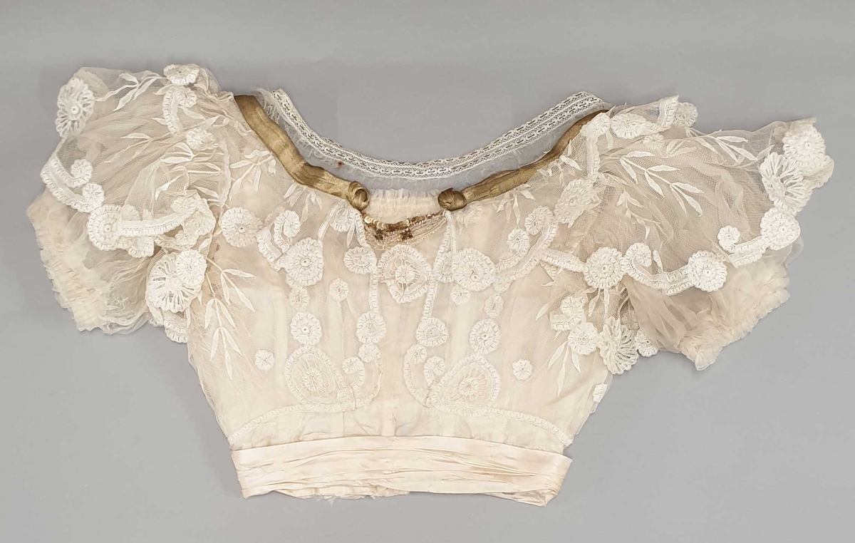 Kjolen av to deler - skjørt med påkneppet bluse.
Blusen er i organza og tyll med formede rosetter av blonder i metervis, påsydd en og en. Et bredt gullbånd med knute i enden rundt halsutringing. Blusen har korte puffermer, med blonder. Skjørtet har slep og består av tyll med broderte rosetter.