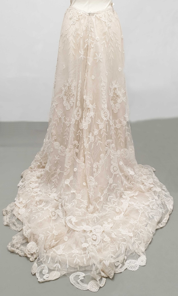 Kjolen av to deler - skjørt med påkneppet bluse.
Blusen er i organza og tyll med formede rosetter av blonder i metervis, påsydd en og en. Et bredt gullbånd med knute i enden rundt halsutringing. Blusen har korte puffermer, med blonder. Skjørtet har slep og består av tyll med broderte rosetter.