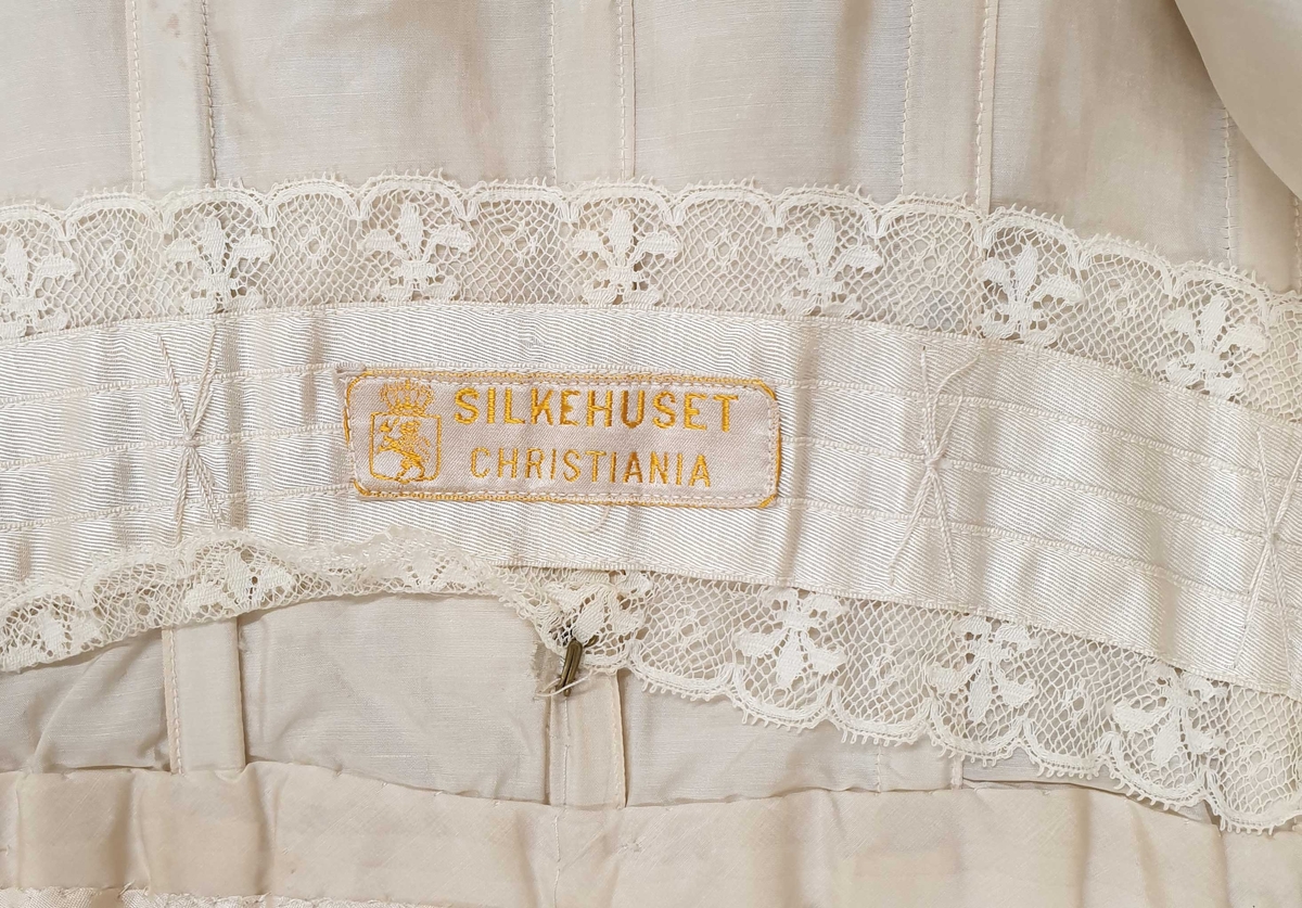 Kjolen av to deler - skjørt med påkneppet bluse.
Blusen er i organza og tyll med formede rosetter av blonder i metervis, påsydd en og en. Et bredt gullbånd med knute i enden rundt halsutringing. Blusen har korte puffermer, med blonder. Skjørtet har slep og består av tyll med broderte rosetter.