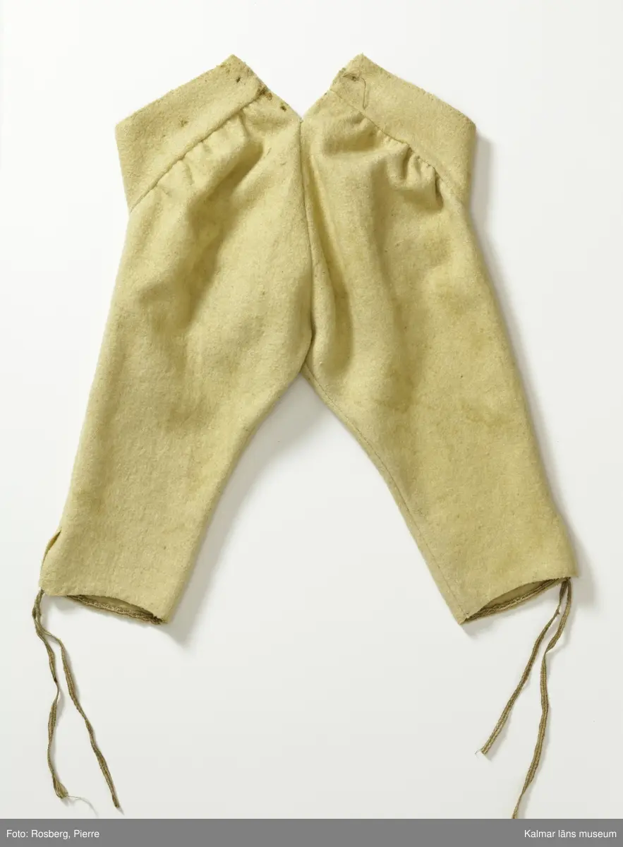 KLM 14854:1. Byxor, knäbyxor. För trumslagare. Kalmar regemente. 1700-talets slut. Korta byxor vävda av gult, troligen bleknat, ylle. Midjegjord/linning knäpps med tre flata gulmetallknappar med ögla fästa på läderrem utanpå fodret. Luckan knäpps med två separata likadana knappar. Sprund mitt bak, två snörhål, rem saknas. Linningens och luckans kanter är fodrade med grovt oblekt linne. Sprund i benslut, knyts med smalt vävt band av grovt garn, linne, fastsytt på insidan så att bara ändarna syns.