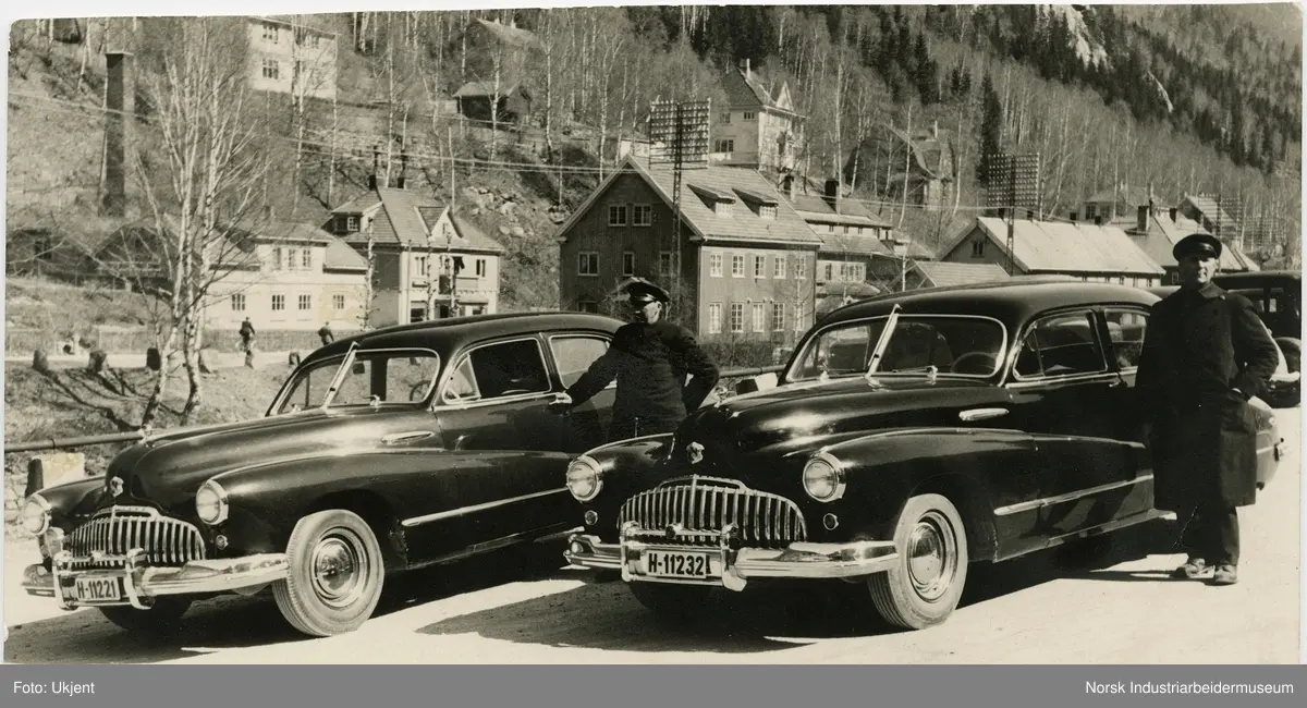 Oskar Andresen og Ole Brenden i uniform stående ved siden av hver sin bil av merket Buick 1946 modell. Bilene tilhører Rjukan Byanlegg og mennene var ansatt som sjåfører i Norsk Hydro. 
Rjukan Varelager, slakteri og bakeri i bakgrunnen. Boliger i Villaveien.