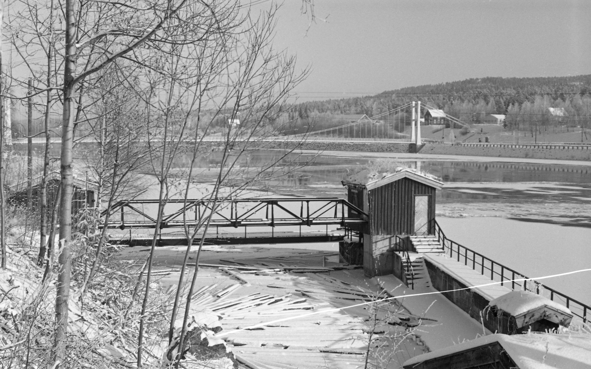 Fra inntakssona til tømmerrenna ved Rånåsfoss i Glomma, i Nes kommune i Akershus, i 1955. Fotografiet er tatt en vinterdag med tilløp til isskorpe på kraftverksdammen. I innløpet til renna lå det en del fastfrosne tømmerstokker med litt snø på. Over inntaksrenna gikk det ei stålbru, og på hver side var det ei lita hytte med saltak, muligens over de mekanismene som ble brukt til å justere posisjonen til «inntaksskuffen», ei kraftig strålrenne som befant seg under vannspeilet eller isflata, og som ble brukt til å regulere gjennomstrømminga gjennom dammen med. Ved Rånåsfossen skal denne skuffen ha vært av noe som ble kalt «Gran-Meyers patenterte konstruksjon». Rånåsfoss kraftstasjon ble bygd for Akershus fylke i perioden 1918-1922. Inntaksmagasinet har et areal på cirkla 10 kvadratkilometer og strekker seg drøyt 2 mil oppover i elveløpet. Dammen fikk et isløp nærmest kraftstasjonen, deretter to 50 meter brede sektorløp for fløtingstømmeret og et 45 meter bredt valseløp mot den vestre elvebredden. Valsen ble brukt til flomregulering og ved tapping vinterstid. Gjennom de store sektorlukene skulle det være mulig å ekspedere 700 000 tømmerstokker i døgnet med et beskjedent tap av vann. I tillegg ble det bygd ei 800 meter lang tømmerrenne av betong på den vestre elvebredden. Den skulle fortrinnsvis brukes når vannføringa var liten. I slike perioder skulle det være mulig å ekspedere 180 000 stokker i døgnet gjennom renna med et vannforbruk som ikke oversteg 30 kubikkmeter i sekundet. På grunn av strømforholdene viste det seg at «påstikningsarbeidet» - det å få tømmeret inn i renna fra damsida - ble mannskapskrevende. Derfor var det bare i ekstreme tørkesomrer at renna var i bruk - ellers gikk tømmeret over sektorløpene. I bakgrunnen på dette fotografiet ser vi Rånåsfoss bru, ei hengebru som ble tegnet og kalkulert av vegingeniøren Olaf Stang (1871-1956) og sto ferdigbygd i 1927. Med en avstand på drøyt 183 meter mellom de to tårnene var dette da et av Norges lengste bruspenn.