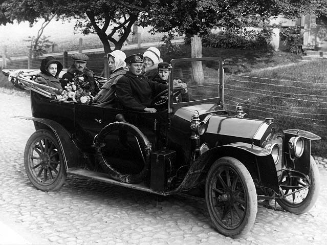 En bil (cabriolet) med registreringsskylt står på Brahegatan i Gränna, vid torgets södra uppfart, riktning söderut. I bilen sitter två män, tre kvinnor och en pojk. Den senare sitter fram vid en man som bär liknande chaufförsuniform.
Någon av personerna är Marsiman från London. Mannen i baksätet är troligen doktor Eurén från Jönköping och någon av kvinnor är fröken Eurén.