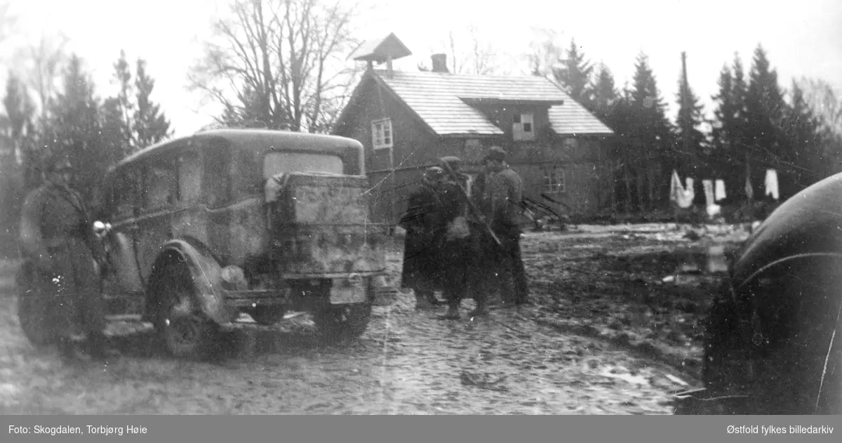 Fra gården Salmonrud i Eidsberg  i april 1940, kaptein Rolf Graffs batteri under tilbaketrekning fra Fossum. Personbil og mennesker på gjørmete gårdstun. I bakgrunnen våningshus.