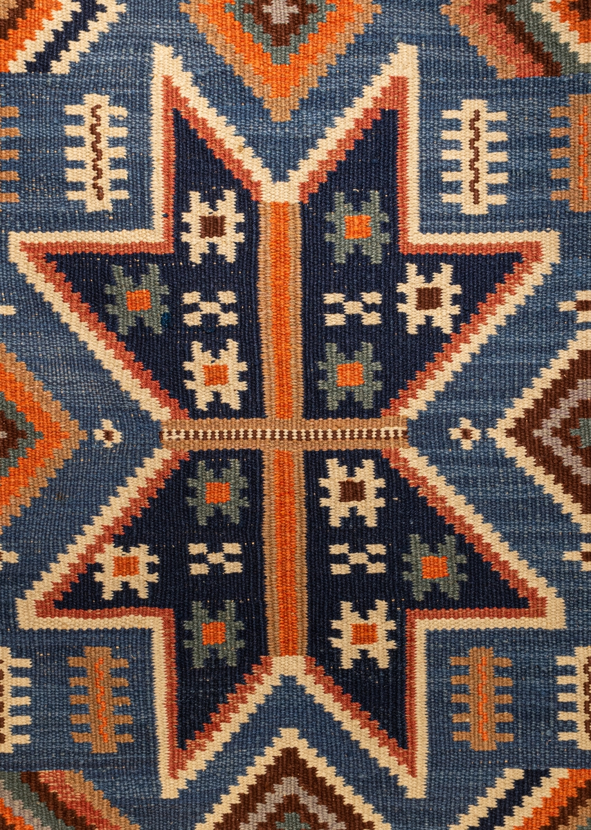 Matta 301 x 232 cm. "Nio stjärnor". Röllakan. Varp av lingarn Inslag av ullgarn. Blå botten med nio riddarstjärnor i flera färger. rik bård med T-ränder och zigzagrand. Formgiven av Märta Måås-Fjetterström under hennes tid som vävlärare vid Vävskolan i Vittsjö, som drevs av Lille Zickerman i regi av Svensk Hemslöjd. Sign. MMF. Köpt 1916-1917 för Gustaf Bergströms gästrum på Hofors, ärvd av Hedvig Ulfsparre vid Bergströms död.