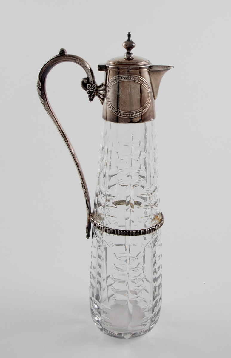 Høyreist kanne i klart glass med påmontert hank og lokk i sølvplett. Konisk korpus som er dekorert med sliping, i form av vertikale linjer som flankeres av parvis olivensliping. Svungen hank i sølvplett med blomsterdekor. Plettmonteringen på kannens øvre del har graverte ovaler, samt innpunslet inskripsjon: "Erindring / af / 23.9.82". Mykt buet nebb samt henglset lokk med balusterformet lokknapp.