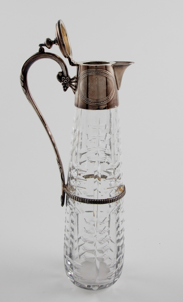 Høyreist kanne i klart glass med påmontert hank og lokk i sølvplett. Konisk korpus som er dekorert med sliping, i form av vertikale linjer som flankeres av parvis olivensliping. Svungen hank i sølvplett med blomsterdekor. Plettmonteringen på kannens øvre del har graverte ovaler, samt innpunslet inskripsjon: "Erindring / af / 23.9.82". Mykt buet nebb samt henglset lokk med balusterformet lokknapp.
