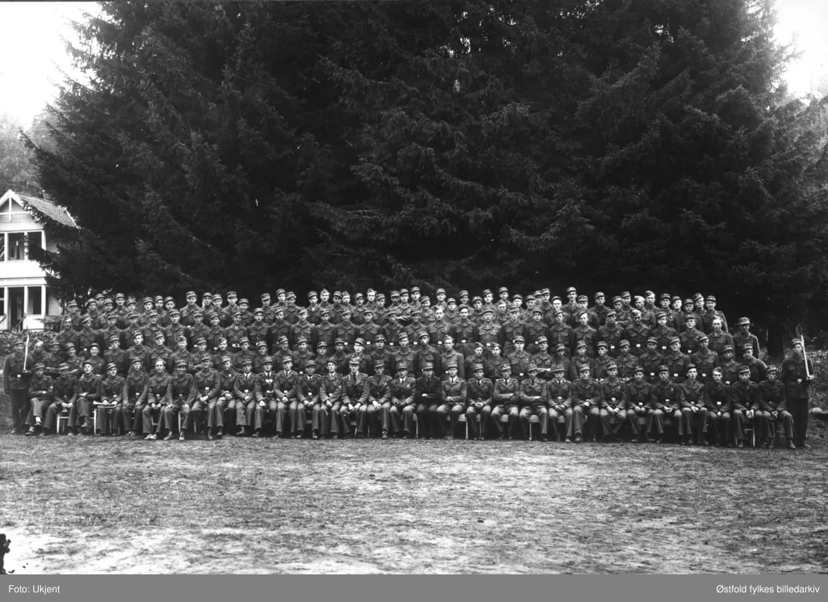 Arbeidstjenesten (AT) under andre verdenskrig, gruppebilde med mannskap og offiserer, utenfor Høysand badehotell i Skjeberg 1942. 
En person er kjent, plassering se fotokort med pålimt utsnitt med kryss under person i første rekke: Sveitfører Grønnerød.