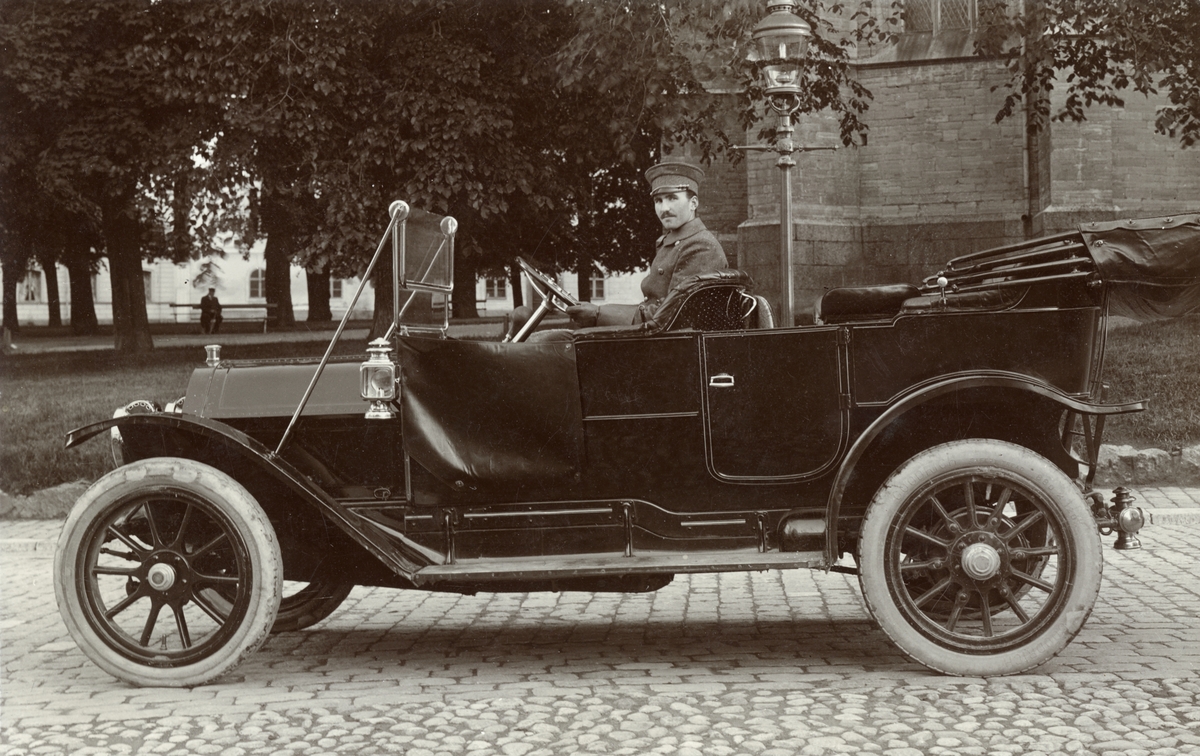 Chauffören Henrik Alm sitter stolt i en Cadillac modell 30 från 1911. Bilen tillhör manufakturhandeln Axel Karlsson & Co där Alm varit anställd sedan omkring 1915. Vid fototillfället har bilen parkerats på Ågatan intill Domkyrkoparken.