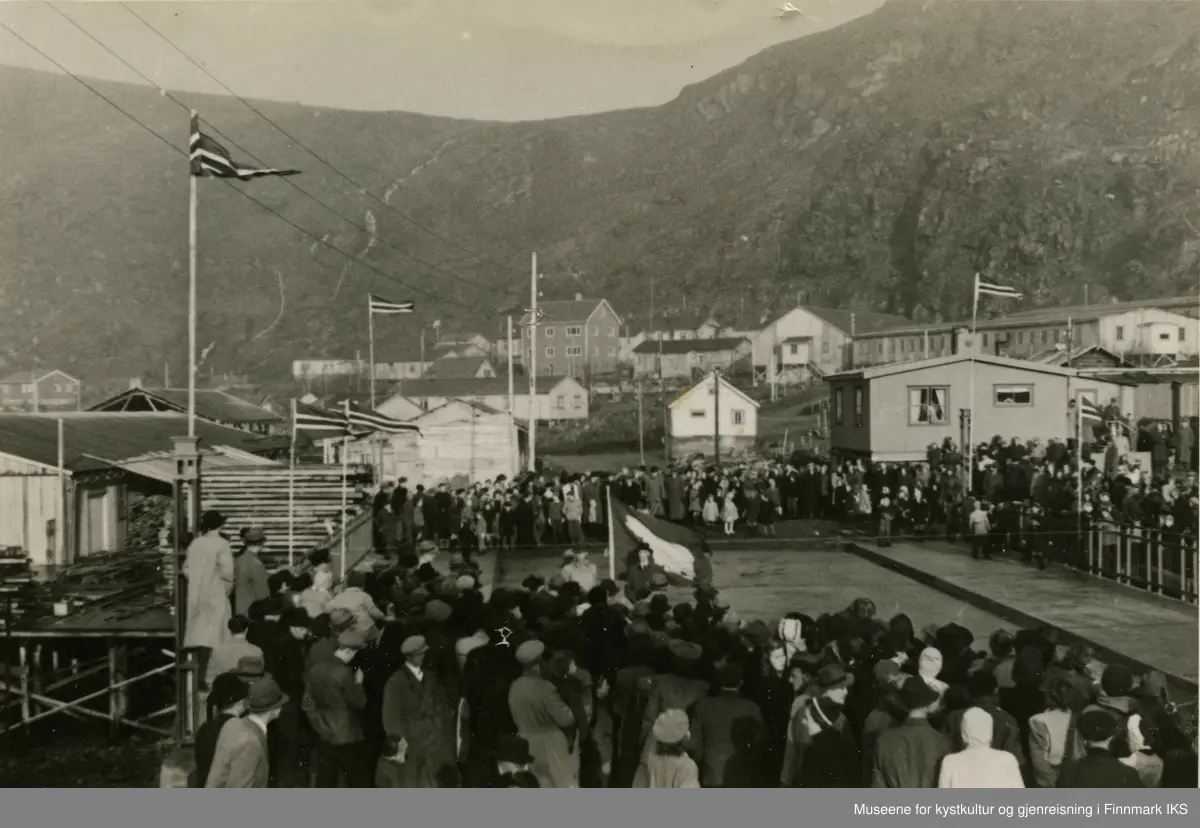 Folk er forsamlet på begge sider av brua over storelva i Hammerfest. I midten av bildet vaier isbjørnflagget og det ser ut som om det holdes en tale. Til venstre ser man Hauans materialhandel. Bebyggelsen er fortsatt preget av brakker, dog har enkelte gjenreisnigshus kommet opp. Oppe til venstre ser man strømledningen. I bakgrunn ser man mollafjellet med mollabekken.