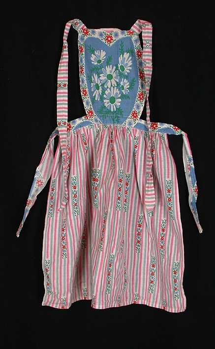 Barnförkläde med bröstlapp. Vitt bomullstyg med lodräta grå och rosa ränder med partier av blommor och blad. Bröstlappen blå och vit med blommor och blad i rött, blått och grönt. Band att knyta runt midja och nacke.