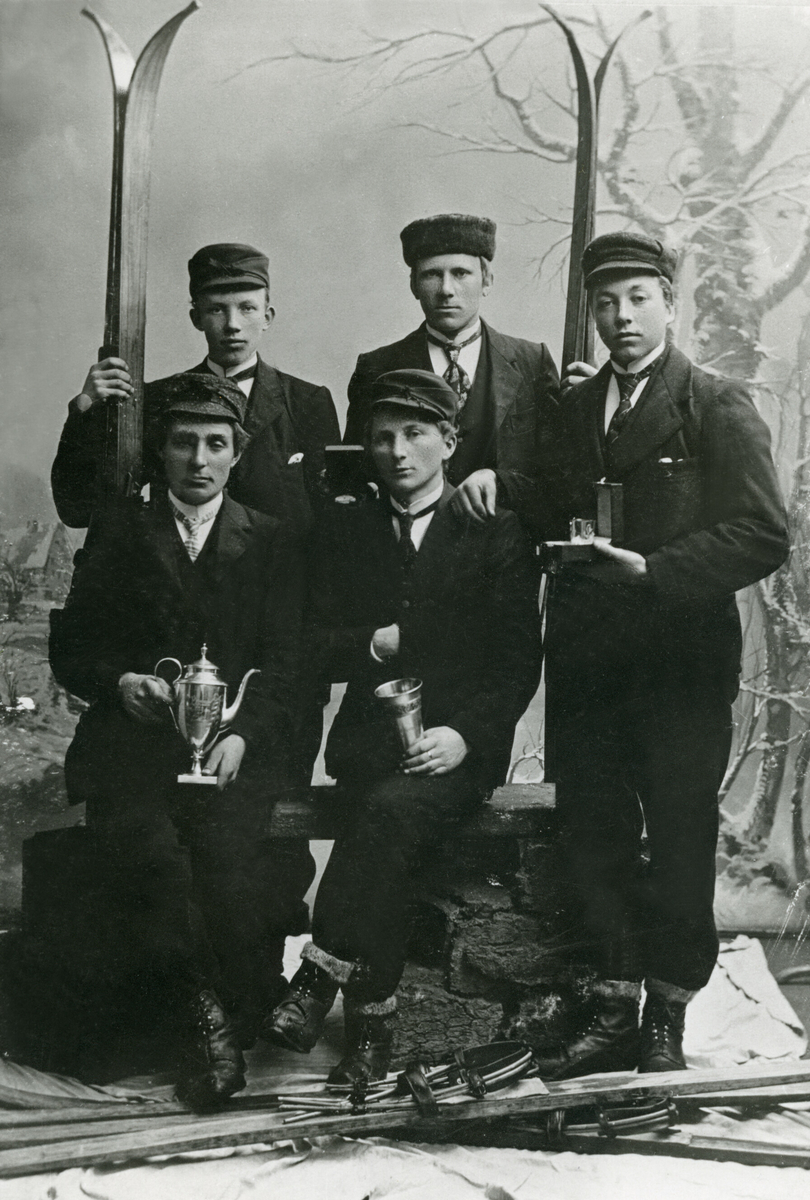 5 yngre menn med ski og premiar fotograferte i atelier. Bildet er teke i etterkant av eit landsrenn i Kikutbakken i Skien i 1904. 
Framme frå v. sit: Olav A. Tjønna og Sveinung G. Bø.
Bak frå v. stend: Olav O. Ajer, Gregar G. Bø og Sveinung G. Kåsa. 
Sveinung G. Bø sit med kongepokalen.