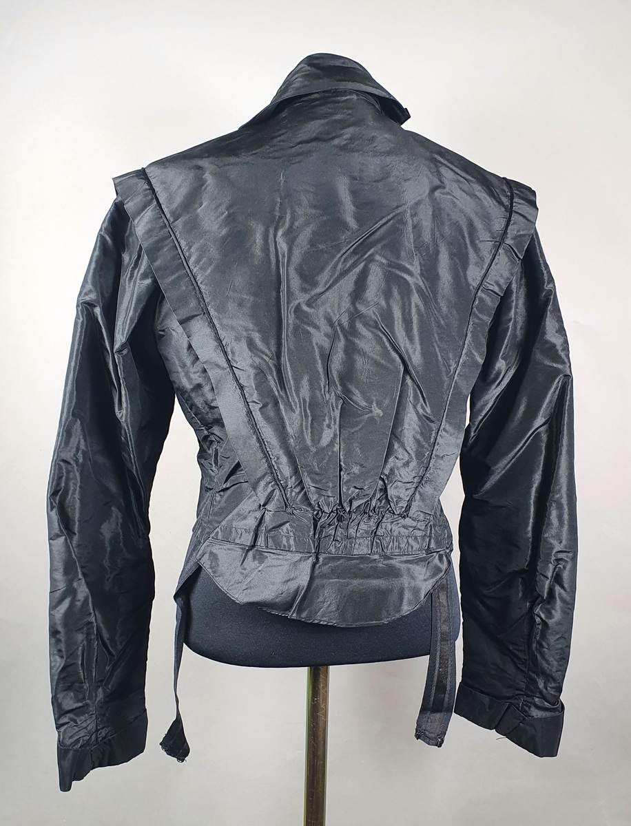 Kjole bestående av to deler - kjoleliv og skjørt. Kjolelivet har dobbelt stoff over rygg og byste, med skjortekrage og slag. Lukkes foran med trykknapper under pynteknapper på det underste laget. Fôret med lin. Svart skjørt med sløyfe på ryggen. Lukkes i ryggen med trykknapper. Stor vidde på skjørtet.