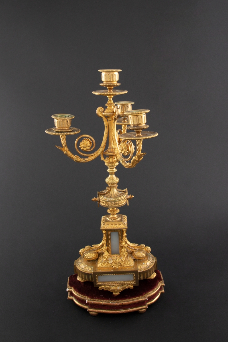 Forgylt kandelaber av bronse med rik dekor i Louis XVI stil. Den er lagd for fire talglys: én som forlengelse av skaft og på tre svungne armer. Skaftets midtre er formet som en urne som står på en rikt dekorert base med innfelt gråhvit alabast. Kandelaberet står på en tresokkel trukket med vinrød fløyel og med forgylte lister og kuleben. Sidene er buet.