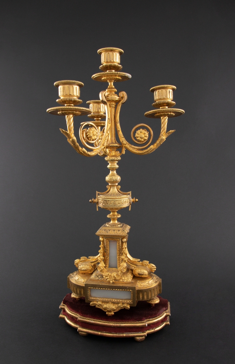 Forgylt kandelaber av bronse med rik dekor i Louis XVI stil. Den er lagd for fire talglys: én som forlengelse av skaft og på tre svungne armer. Skaftets midtre er formet som en urne som står på en rikt dekorert base med innfelt gråhvit alabast. Kandelaberet står på en tresokkel trukket med vinrød fløyel og med forgylte lister og kuleben. Sidene er buet.
