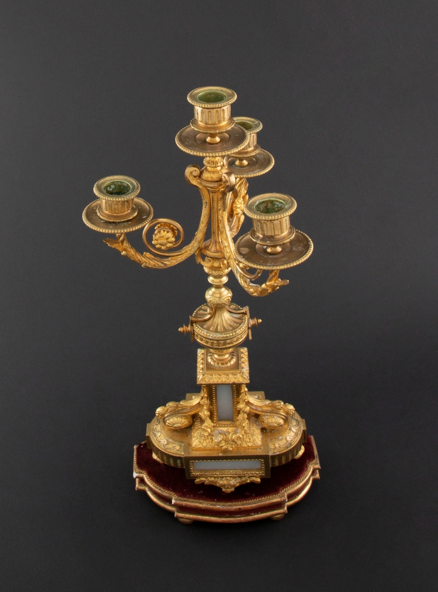 Forgylt kandelaber av bronse med rik dekor i Louis XVI stil. Den er lagd for fire talglys: én som forlengelse av skaft og på tre svungne armer. Skaftets midtre er formet som en urne som står på en rikt dekorert base med innfelt gråhvit alabast. Kandelaberet står på en tresokkel trukket med vinrød fløyel og med forgylte lister og kuleben. Sidene er buet.