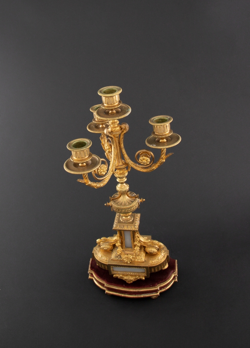 Forgylt kandelaber av bronse med rik dekor i Louis XVI stil. Den er lagd for fire talglys: én som forlengelse av skaft og på tre svungne armer. Skaftets midtre er formet som en urne som står på en rikt dekorert base med innfelt gråhvit alabast. Kandelaberet står på en tresokkel trukket med vinrød fløyel og med forgylte lister og kuleben. Sidene er buet.
