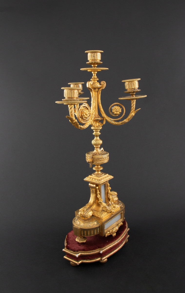 Forgylt kandelaber av bronse med rik dekor i Louis XVI stil. Den er lagd for fire talglys: én som forlengelse av skaft og på tre svungne armer. Skaftets midtre er formet som en urne som står på en rikt dekorert base med innfelt gråhvit alabast. Kandelaberet står på en tresokkel trukket med vinrød fløyel og med forgylte lister og kuleben. Sidene er buet.