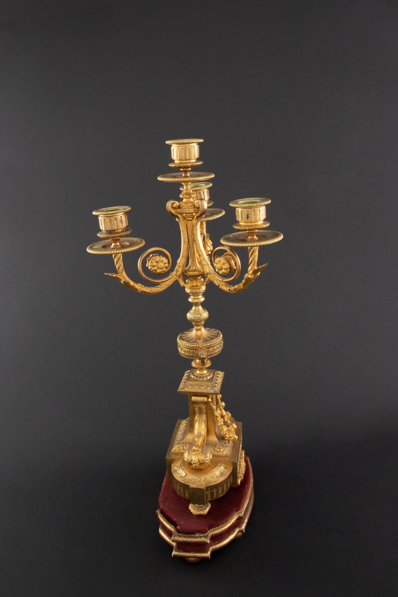 Forgylt kandelaber av bronse med rik dekor i Louis XVI stil. Den er lagd for fire talglys: én som forlengelse av skaft og på tre svungne armer. Skaftets midtre er formet som en urne som står på en rikt dekorert base med innfelt gråhvit alabast. Kandelaberet står på en tresokkel trukket med vinrød fløyel og med forgylte lister og kuleben. Sidene er buet.
