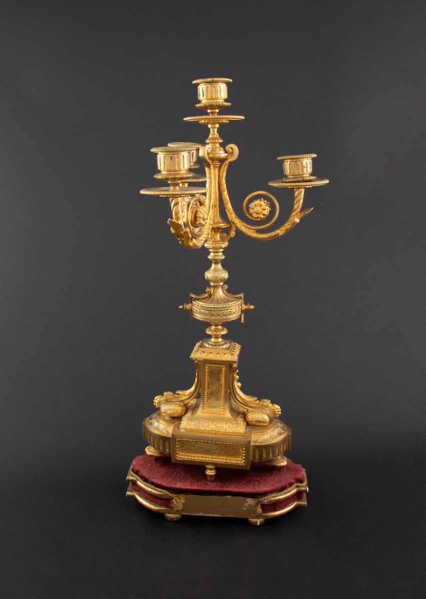 Forgylt kandelaber av bronse med rik dekor i Louis XVI stil. Den er lagd for fire talglys: én som forlengelse av skaft og på tre svungne armer. Skaftets midtre er formet som en urne som står på en rikt dekorert base med innfelt gråhvit alabast. Kandelaberet står på en tresokkel trukket med vinrød fløyel og med forgylte lister og kuleben. Sidene er buet.