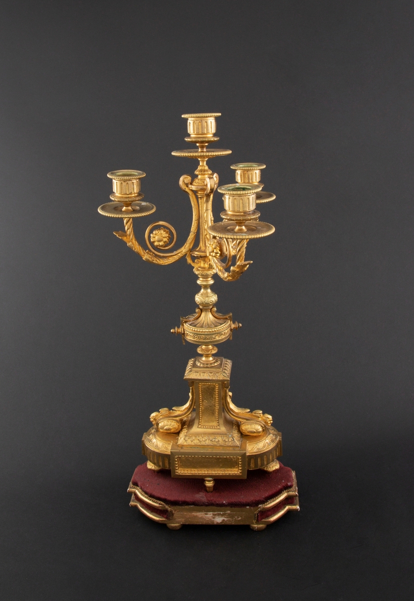 Forgylt kandelaber av bronse med rik dekor i Louis XVI stil. Den er lagd for fire talglys: én som forlengelse av skaft og på tre svungne armer. Skaftets midtre er formet som en urne som står på en rikt dekorert base med innfelt gråhvit alabast. Kandelaberet står på en tresokkel trukket med vinrød fløyel og med forgylte lister og kuleben. Sidene er buet.
