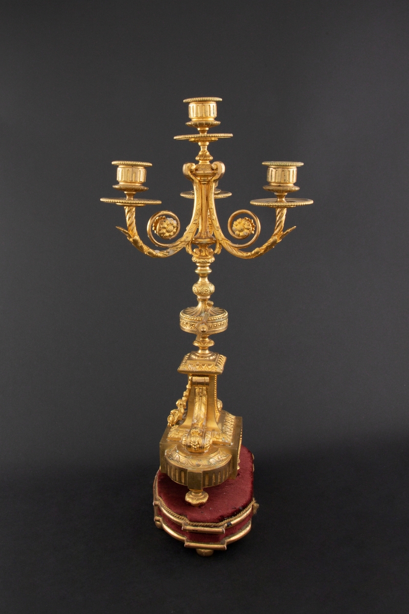 Forgylt kandelaber av bronse med rik dekor i Louis XVI stil. Den er lagd for fire talglys: én som forlengelse av skaft og på tre svungne armer. Skaftets midtre er formet som en urne som står på en rikt dekorert base med innfelt gråhvit alabast. Kandelaberet står på en tresokkel trukket med vinrød fløyel og med forgylte lister og kuleben. Sidene er buet.