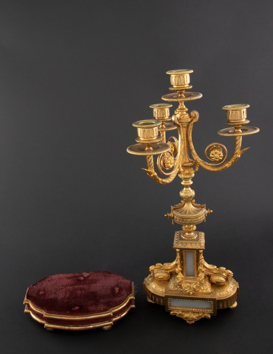 Forgylt kandelaber av bronse med rik dekor i Louis XVI stil. Den er lagd for fire talglys: én som forlengelse av skaft og på tre svungne armer. Skaftets midtre er formet som en urne som står på en rikt dekorert base med innfelt gråhvit alabast. Kandelaberet står på en tresokkel trukket med vinrød fløyel og med forgylte lister og kuleben. Sidene er buet.
