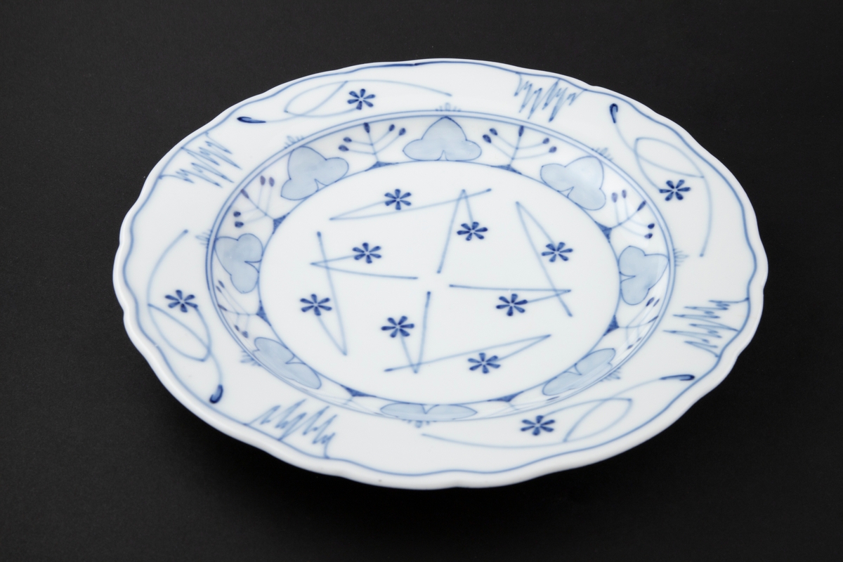 Sirkulær dyp tallerken i porselen dekorert med koboltblå underglasurmaling. Bunnen er dekorert med åtte blåveisblomster, omgitt av geometriske streker som danner et svastikaligende mønster. Den er omkranset av en sirkulær bord bestående treflikede blåveisblader samt flerarmede stiliserte plantemotiver. Langs tallerkenens fane gjenopptas blåveismotivet, omgitt av slyngende linjeføringer samt fire stiliserte istapper. Tallerkenen er uten stempel.
