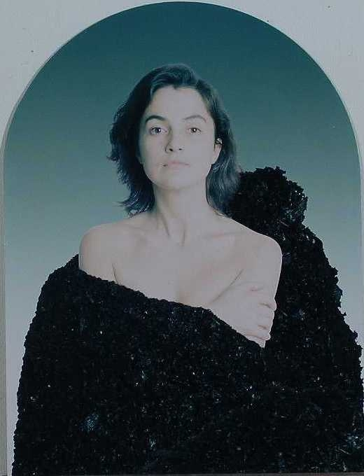 Midjebild av person med axellångt, vågigt mörkt hår. Kroppen draperad i svart tyg, delar av överkroppen blottad.