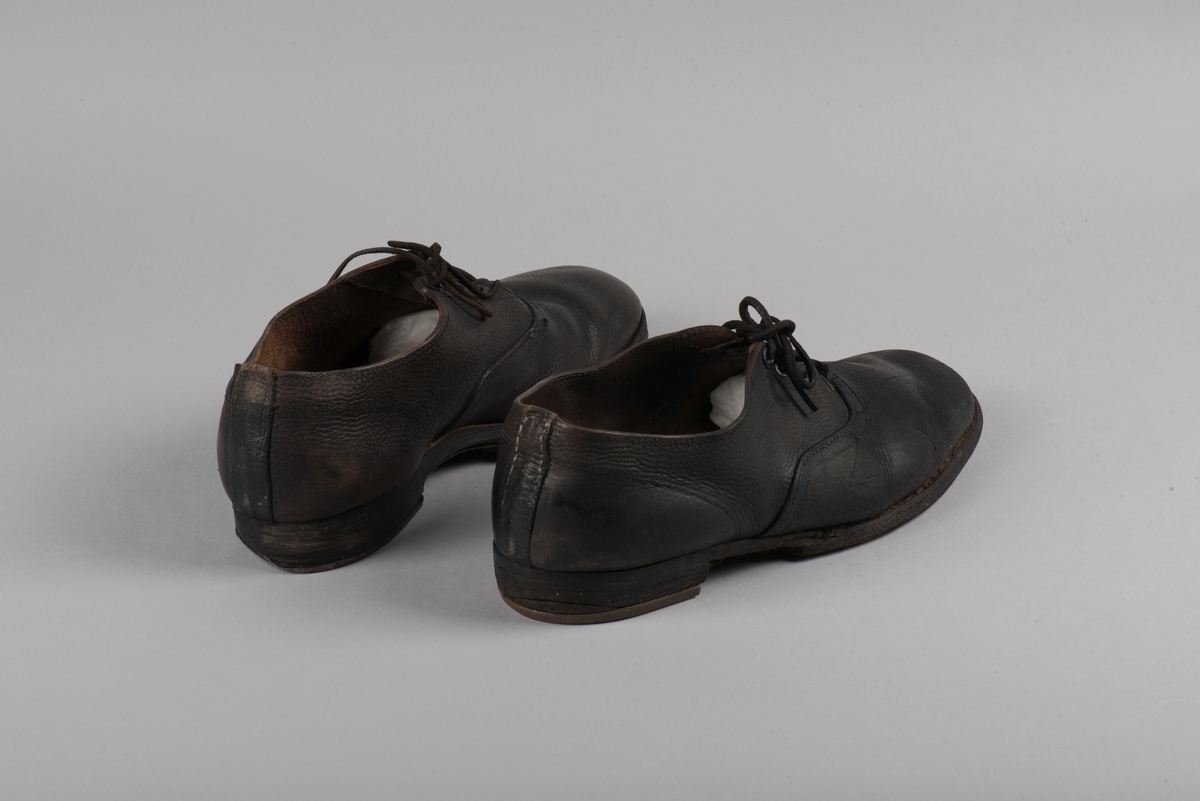 Herrsko, ett par, av skinn (läder) med sula av läder. Klacken skodd med metallsko i U-form. fyra par hål med öljetter i metall och lädersnöre. Ofodrade. Möjligen storlek 46 eller 40. Skinnet är präglat med ett möster av små stjärnor. 

På den högra skon är metallskoningen på klacken vänd, troligen omspikad. 

Skorna hör till en uppsättning kläder för en manlig fånge. 
Kläderna består av en kort dubbelknäppt rock, ett par byxor, en väst av grå vadmal, en mössa och en skjorta av oblekt linne.