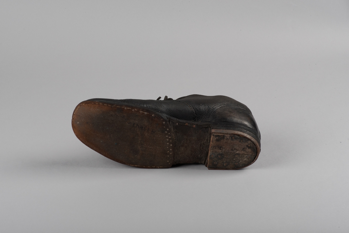 Herrsko, ett par, av skinn (läder) med sula av läder. Klacken skodd med metallsko i U-form. fyra par hål med öljetter i metall och lädersnöre. Ofodrade. Möjligen storlek 46 eller 40. Skinnet är präglat med ett möster av små stjärnor. 

På den högra skon är metallskoningen på klacken vänd, troligen omspikad. 

Skorna hör till en uppsättning kläder för en manlig fånge. 
Kläderna består av en kort dubbelknäppt rock, ett par byxor, en väst av grå vadmal, en mössa och en skjorta av oblekt linne.