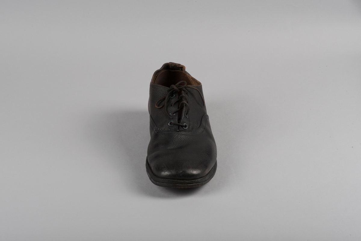 Herrsko, ett par, av skinn (läder) med sula av läder. Klacken skodd med metallsko i U-form. fyra par hål med öljetter i metall och lädersnöre. Ofodrade. Möjligen storlek 46 eller 40. Skinnet är präglat med ett möster av små stjärnor. 

På den högra skon är metallskoningen på klacken vänd, troligen omspikad. 

Skorna hör till en uppsättning kläder för en manlig fånge. 
Kläderna består av en kort dubbelknäppt rock, ett par byxor, en väst av grå vadmal, en mössa och en skjorta av oblekt linne.