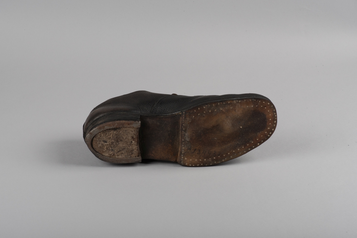 Herrsko, ett par, av skinn (läder) med sula av läder. Klacken skodd med metallsko i U-form. fyra par hål med öljetter i metall och lädersnöre. Ofodrade. Möjligen storlek 46 eller 40. Skinnet är präglat med ett möster av små stjärnor. 

På den högra skon är metallskoningen på klacken vänd, troligen omspikad. 

Skorna hör till en uppsättning kläder för en manlig fånge. 
Kläderna består av en kort dubbelknäppt rock, ett par byxor, en väst av grå vadmal, en mössa och en skjorta av oblekt linne.