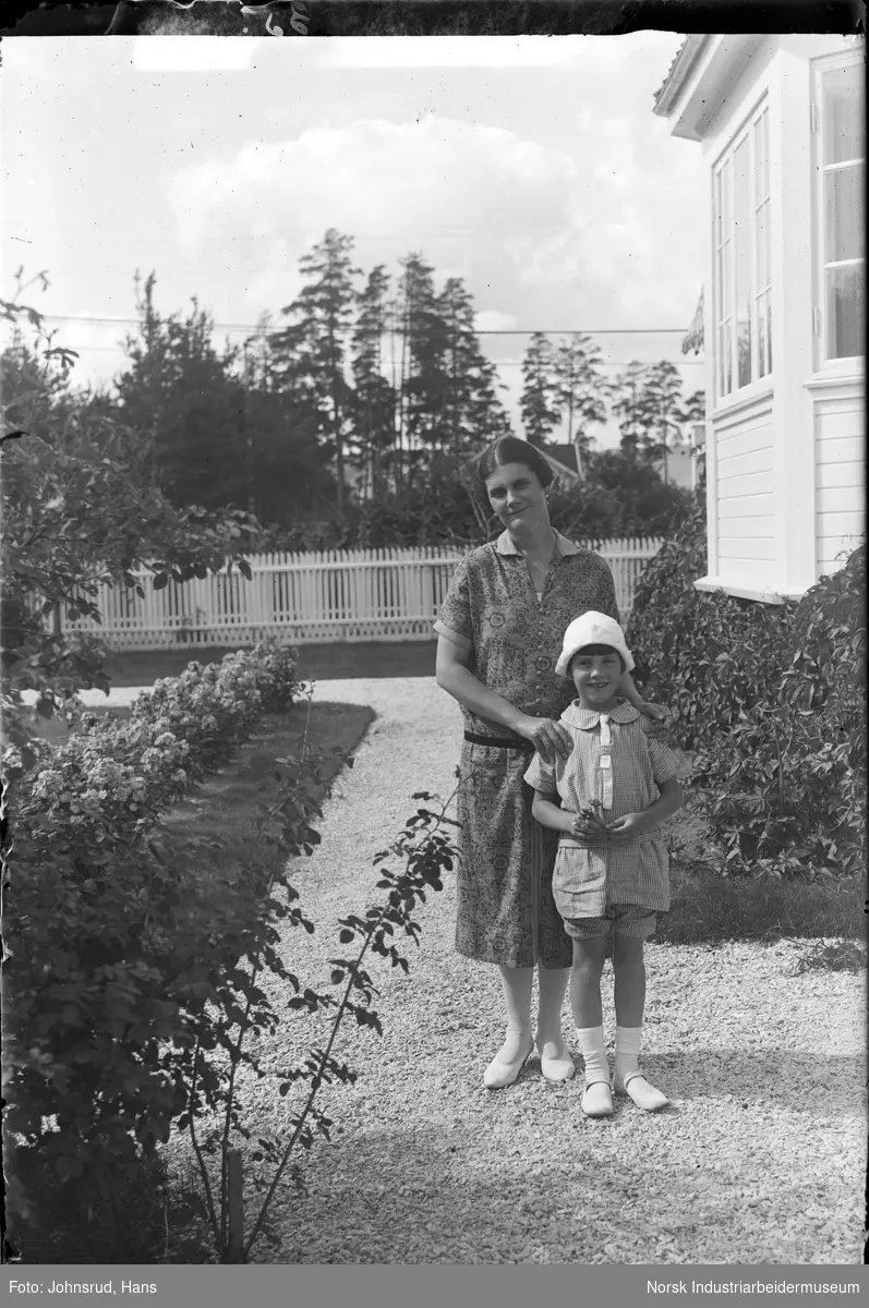 Kvinne med barn stående i hage. Blomster langs gruslagt vei. 
Fotografi bestilt av Dr. Blick.