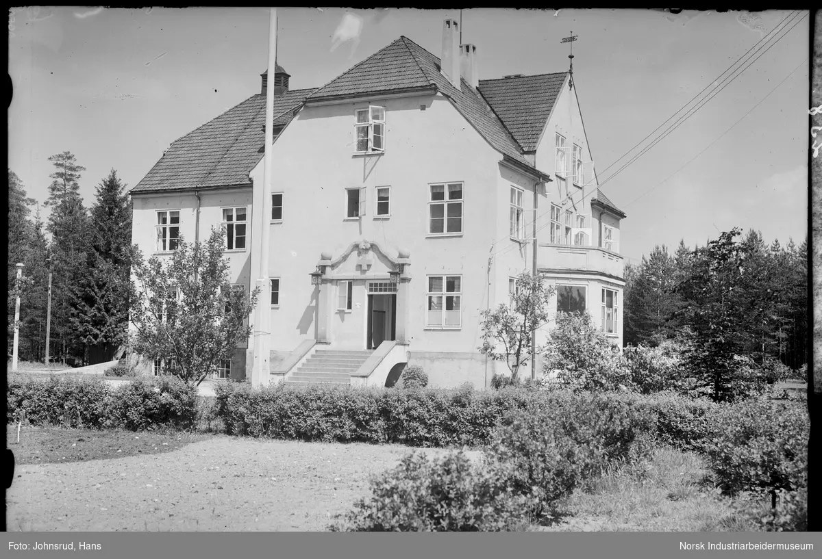 Eksteriør Notodden sykehus. Sykepleier stående delvis gjemt bak busker i hagen.