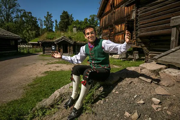 Man in traditional Norwegian folk costume sitting outdoors among historic timber buildings at Norsk Folkemuseum.