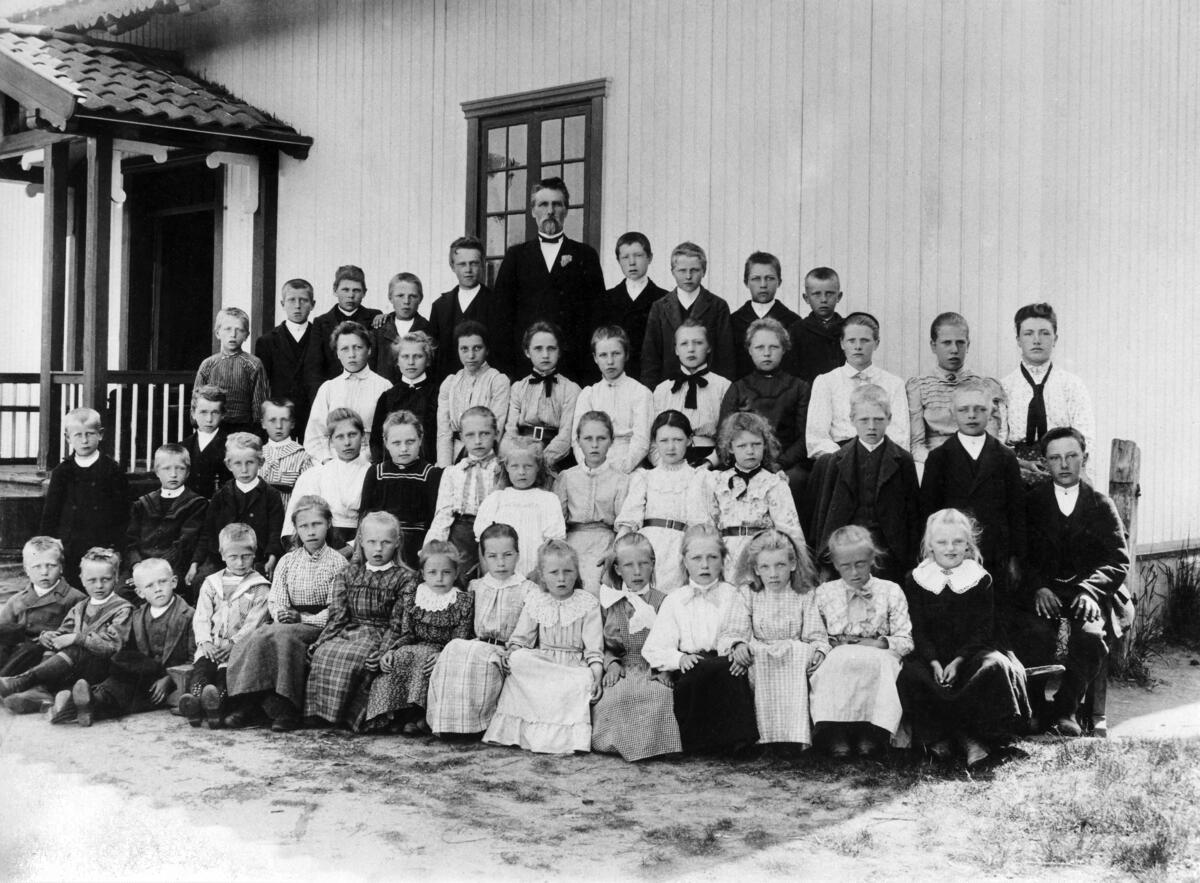 Fotografi i svart-hvitt av en skoleklasse og lærer Simensen. Elevene og læreren blir fotografert utenfor Skinnarbøl skole, som nå er skolemuseum.