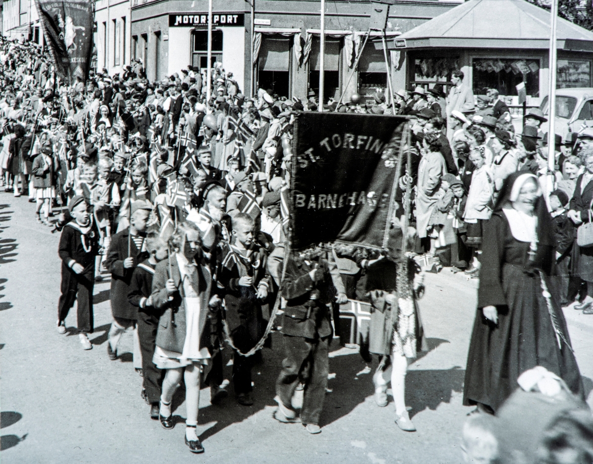 17. mai tog. Tatt utenfor Basarbygningen i Strandgata, nederst på Stortorget med firma Motorsport i hjørnegården i bakgrunnen (Storhamargata 11) Like ovenfor denne gården ser vi Baker Didriksen, derav tilnavnet Kavringbakken på nedre del av Storhamargata. "Skoletoget" kommer ned Storhamargata. Barn fra St. Torfinn barnehage med fanen som har påskrift "St. Torfinns barnehage". Søster Sunniva foran t.h. i bildet.