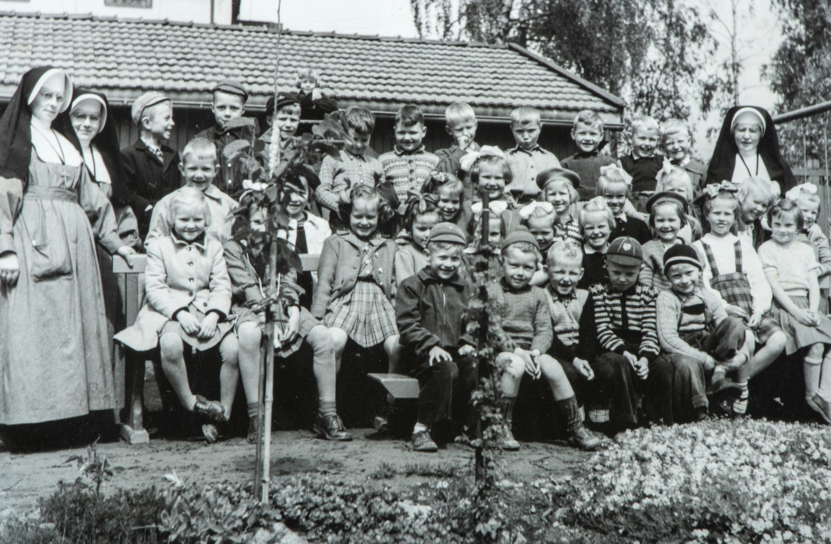Stor gruppe barnehagebarn. St. Torfinn barnehage. Tatt inne i haven til Torggata 113 mot nordøst rundt 1953. 
Arne Hesselberg med kortbukse i fremste rad. Fra v. sr Gudulina og sr. Antona og t.h. sr. Sunniva.