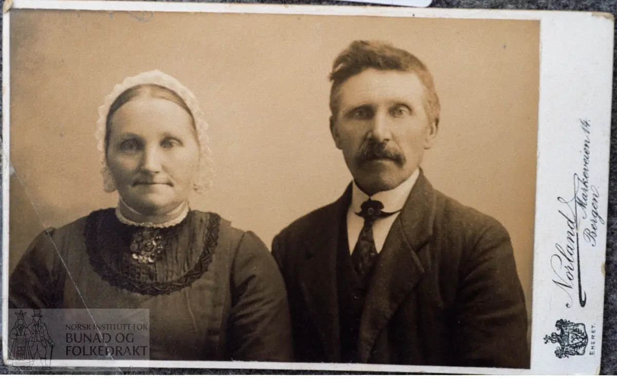 Foto av Henrikka Hallvardtr. Wergeland, f. 1868, og mannen.  Kvitehue.  -  Matr.:  Kvitt bomullslerret med hekla blonde. Knyteband av kvitt bendelband. Trekksnor av smalt bendelband.