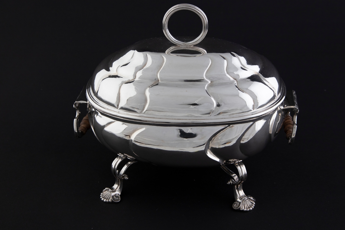 Terrin med lokk i sølv. Den har oval form og står på fire svungne bein som nederst slutter i et muslingmotiv. Korpus og det hvelvede lokket er dekorert med sveifede knekk. På hver kortside er hengslet håndtak med tre-grep. På lokket er det et ringformet grep.