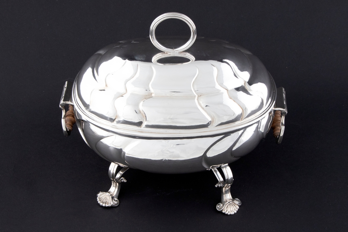 Terrin med lokk i sølv. Den har oval form og står på fire svungne bein som nederst slutter i et muslingmotiv. Korpus og det hvelvede lokket er dekorert med sveifede knekk. På hver kortside er hengslet håndtak med tre-grep. På lokket er det et ringformet grep.