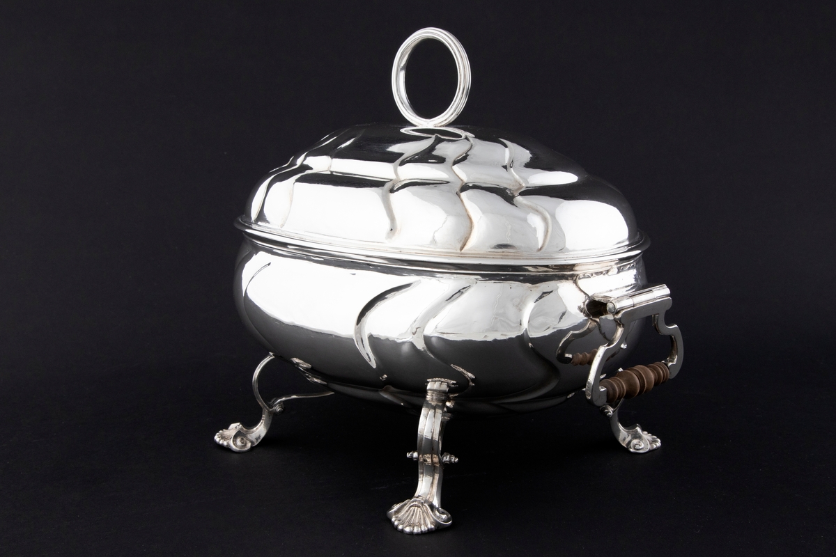 Terrin med lokk i sølv. Den har oval form og står på fire svungne bein som nederst slutter i et muslingmotiv. Korpus og det hvelvede lokket er dekorert med sveifede knekk. På hver kortside er hengslet håndtak med tre-grep. På lokket er det et ringformet grep.