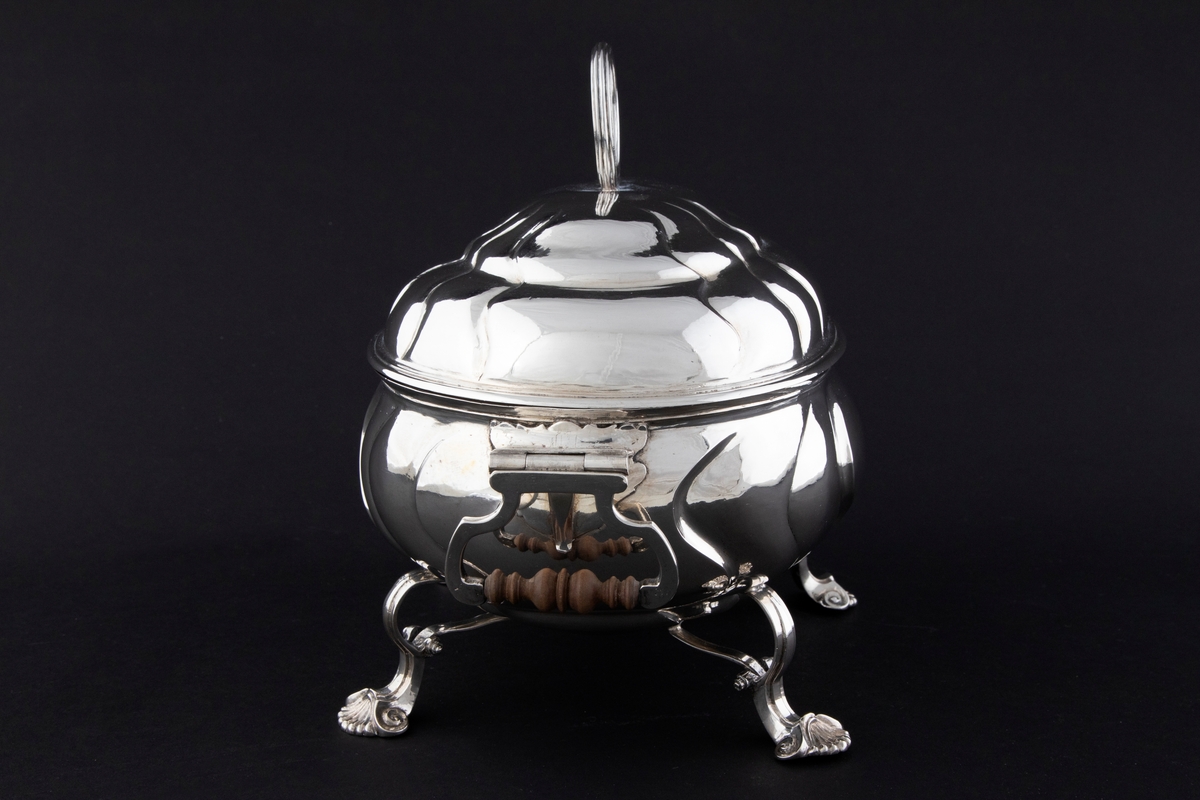 Terrin med lokk i sølv. Den har oval form og står på fire svungne bein som nederst slutter i et muslingmotiv. Korpus og det hvelvede lokket er dekorert med sveifede knekk. På hver kortside er hengslet håndtak med tre-grep. På lokket er det et ringformet grep.