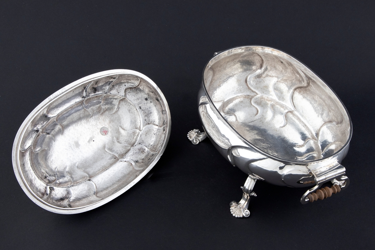 Terrin med lokk i sølv. Den har oval form og står på fire svungne bein som nederst slutter i et muslingmotiv. Korpus og det hvelvede lokket er dekorert med sveifede knekk. På hver kortside er hengslet håndtak med tre-grep. På lokket er det et ringformet grep.
